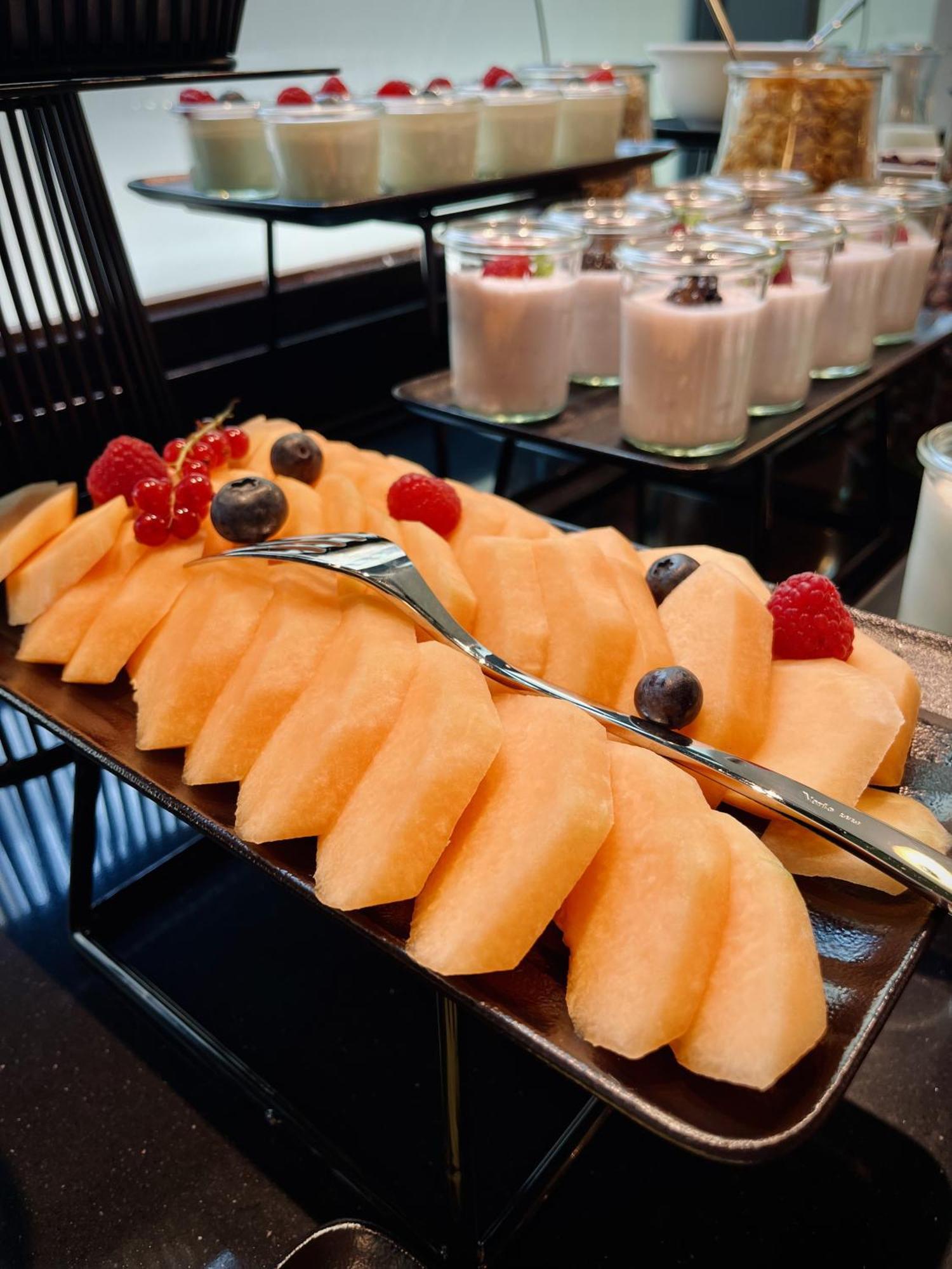 Апарт отель Royal Tulip Warsaw Centre Экстерьер фото The photo features a platter of sliced cantaloupe, arranged neatly and garnished with a few berries. There are red raspberries and blueberries placed on top of the melon slices. In the background, there are cups of dessert, likely some type of puddin