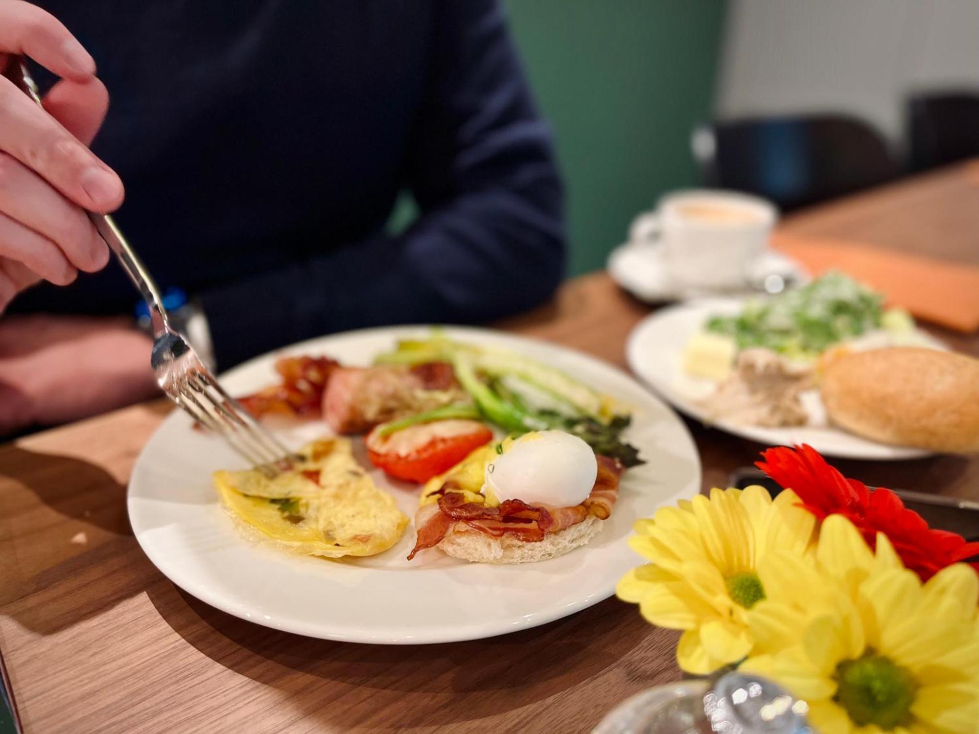 Апарт отель Royal Tulip Warsaw Centre Экстерьер фото The photo displays a breakfast plate with various items. There's an omelet, a poached egg, bacon, sliced tomatoes, and some greens. A hand is reaching for the food with a fork. Additionally, there are flowers on the table and a small cup of coffee. T