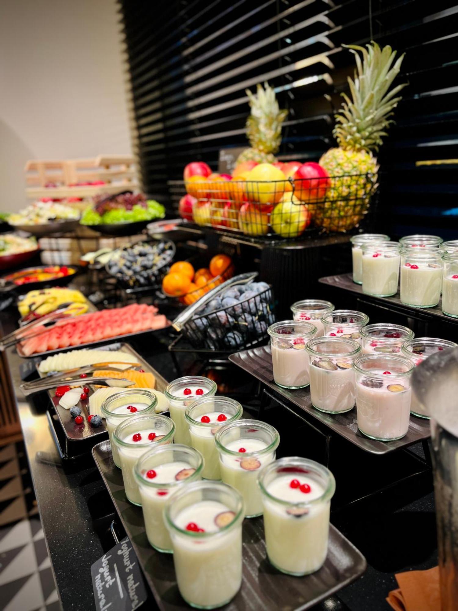 Апарт отель Royal Tulip Warsaw Centre Экстерьер фото The photo displays an assortment of food items arranged on a buffet table. In the foreground, there are several small glass containers filled with creamy desserts, garnished with red berries. Behind them, various fruits are presented, including apple