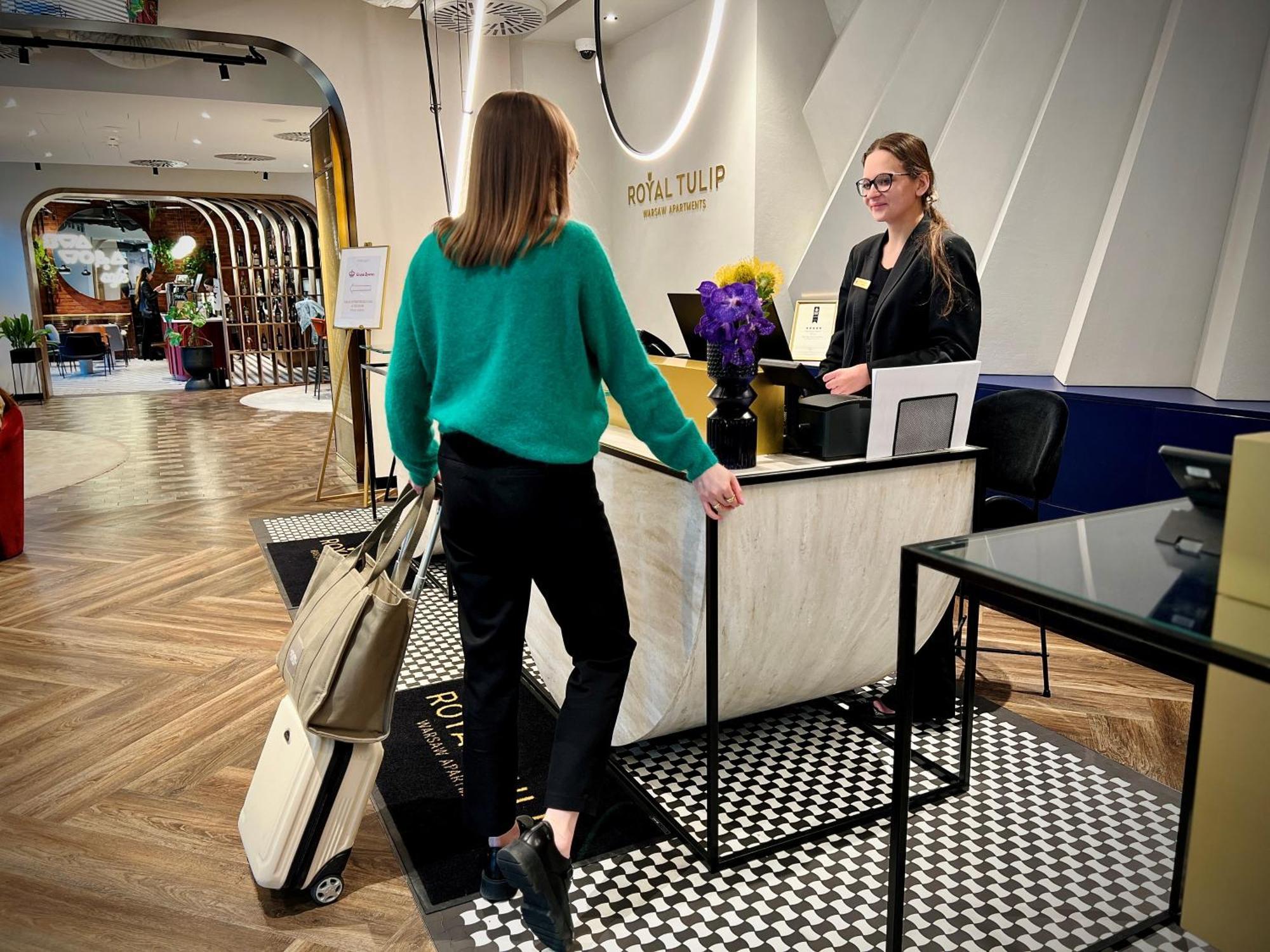 Апарт отель Royal Tulip Warsaw Centre Экстерьер фото The photo depicts a hotel reception area. There is a young woman in a green sweater and black pants approaching the reception desk, carrying a suitcase and a bag. Behind the desk, another woman, likely a hotel staff member, is standing and appears to