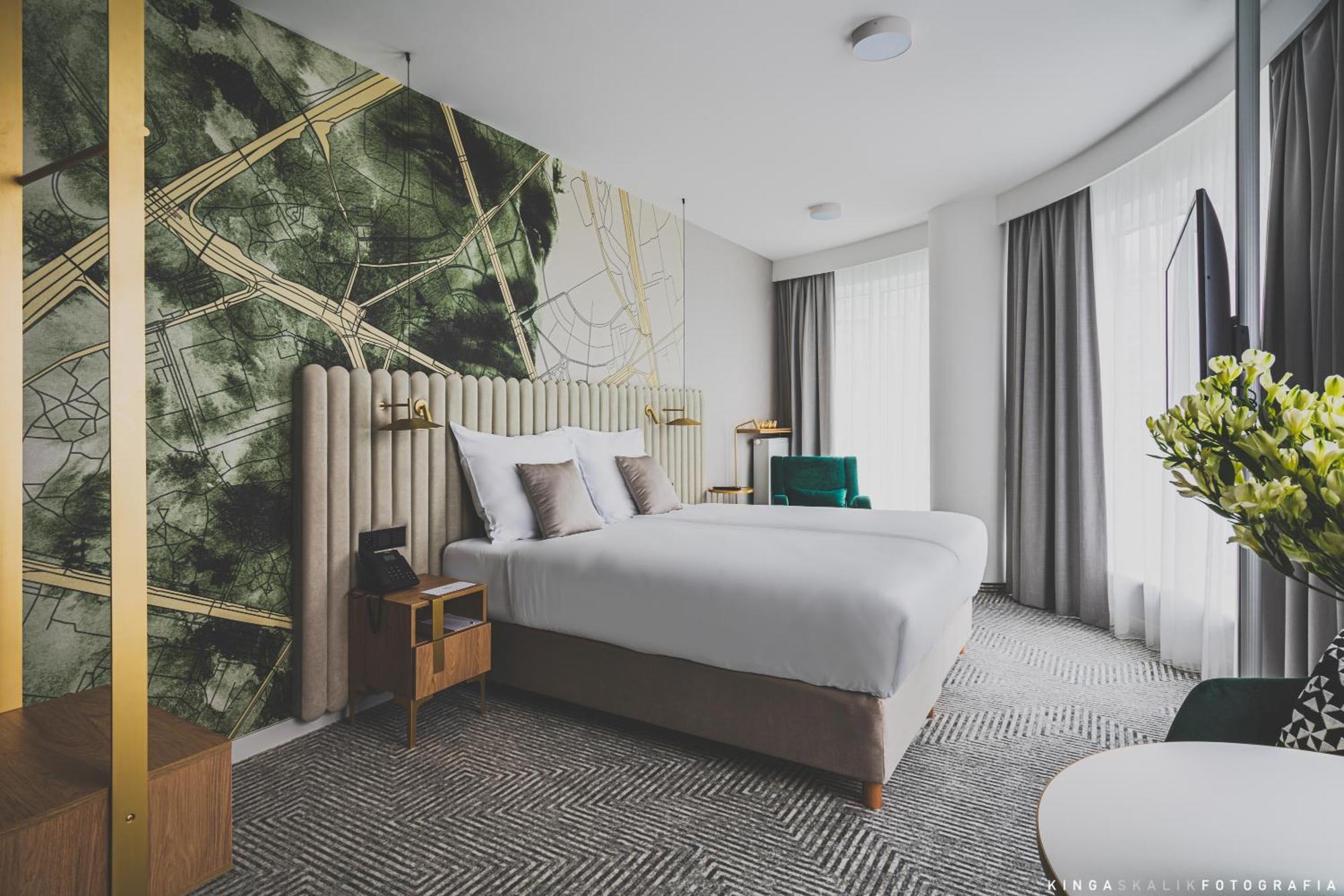 Апарт отель Royal Tulip Warsaw Centre Номер фото The photo shows a modern hotel room featuring two beds with white bedding. The headboard is upholstered and elegantly designed. Behind the beds, there's a striking wall design with abstract green and gold patterns. The room has large windows with lig